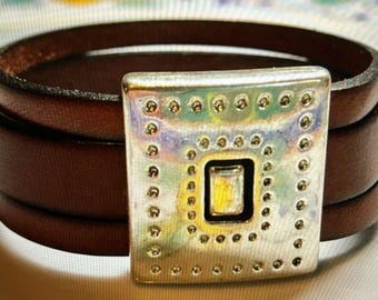 May include: A brown leather wrap bracelet with a silver square buckle featuring a raised, textured design.