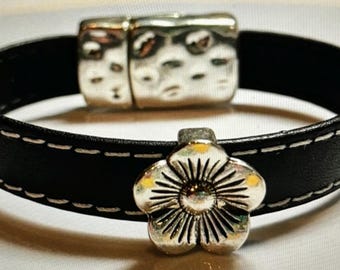 May include: A black leather bracelet with white stitching and a silver flower charm. The bracelet has a magnetic clasp.