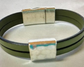 May include: Olive green leather bracelet with a silver magnetic clasp. The bracelet features a double-band design and a rectangular clasp. The metal clasp has a shiny, reflective surface. The bracelet is a piece of jewelry.