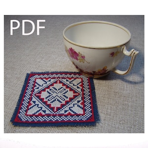 Puede incluir: Un posavasos bordado en azul y rojo con un centro blanco y un patrón geométrico. El posavasos está sobre una superficie de tela beige junto a una taza de té blanca con diseños florales rosas.