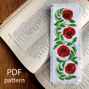 May include: White fabric bookmark with a cross-stitched design of three red poppies with green leaves and vines. The bookmark is placed on an open book with text and the words "PDF pattern" are visible.