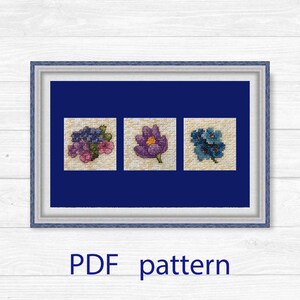 May include: A framed cross-stitch pattern featuring three floral designs. The designs include a cluster of purple and pink flowers, a purple flower, and a blue flower. The frame is blue, and the background is a dark blue. The text "PDF pattern" is at the bottom.