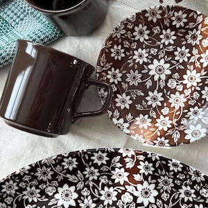 May include: An overhead shot of brown ceramic mugs and plates with floral patterns. The plates have a brown background with white floral designs. The mugs are a solid brown color. The plates and mugs are arranged on a white tablecloth.