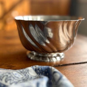 May include: A silver-colored bowl with a fluted design, resting on a small pedestal base. The bowl has a wavy rim and is set on a wooden table. A blue and white patterned cloth is in the foreground.