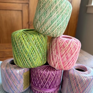May include: A stack of six spools of colorful crochet thread. The spools are in shades of green, pink, purple, and blue. The thread is tightly wound and has a slight sheen. The spools are arranged in a pyramid shape.