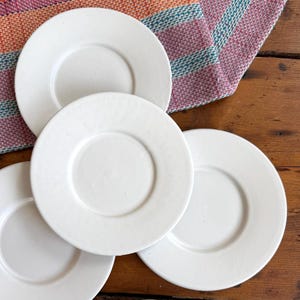 May include: A set of four circular white ceramic saucers with a smooth, matte finish and a slightly raised central indentation. Each saucer features a clean, minimalist design with a flat rim and a consistent, solid off-white color throughout.