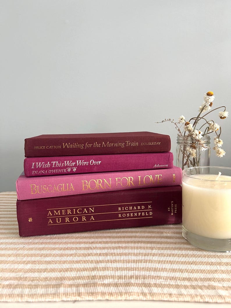 May include: A stack of four books with burgundy, magenta, and pink covers. The titles include "Waiting for the Morning Train," "I Wish This War Were Over," "Born for Love," and "American Aurora." A lit candle in a glass and a small jar of dried flowers are also present.