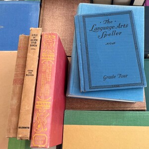 May include: A collection of vintage books with various colors and titles. A blue book titled "The Language Arts Speller" is stacked on top of other books. Other books include "Girls of Silver Spur Ranch" and "Theater Shows".