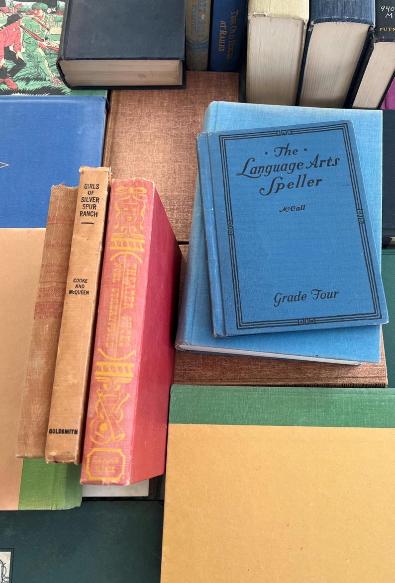 May include: A collection of vintage books with various colors and titles. A blue book titled "The Language Arts Speller" is stacked on top of other books. Other books include "Girls of Silver Spur Ranch" and "Theater Shows".