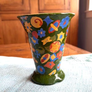May include: A small, flared ceramic vase with a glossy dark green base color. The surface is decorated with a colorful, hand-painted pattern of various shapes, including yellow stars, blue flowers, red circles, and orange abstract geometric motifs.