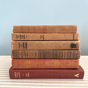 May include: A stack of vintage books with aged covers in shades of brown and red. Titles include "Girls of Silver Spur Ranch," "American Boy Sports Stories," and "Theater From Moel Streatp." The books are stacked on a striped surface.