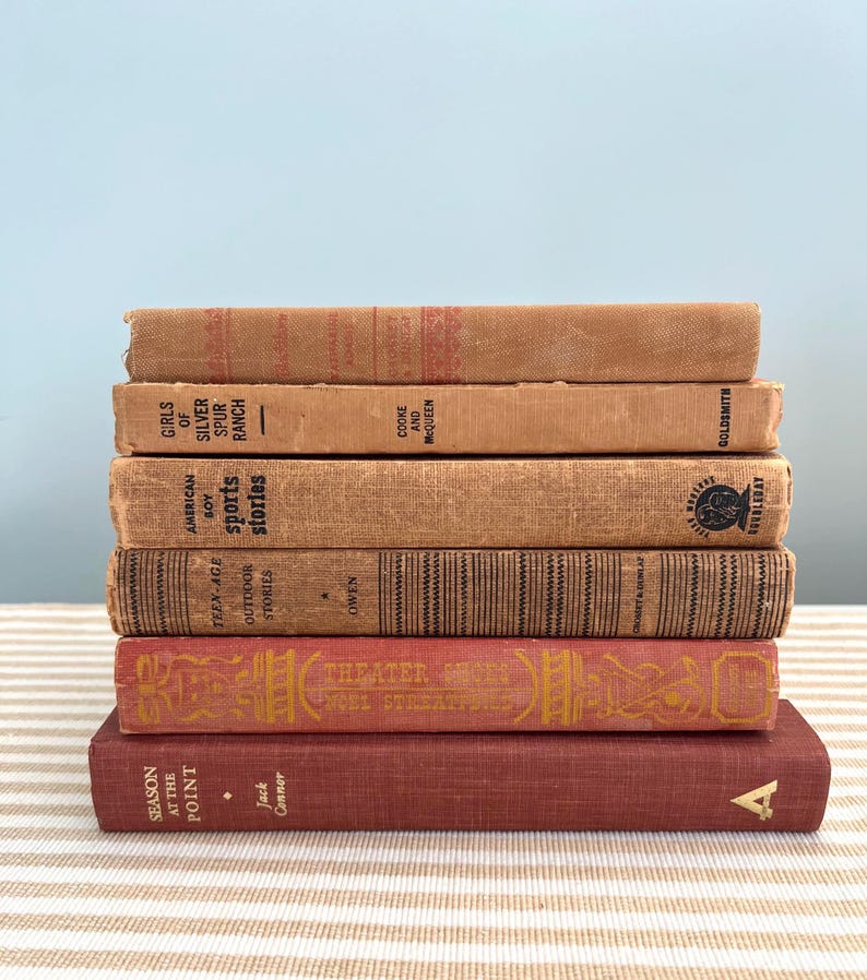 May include: A stack of vintage books with aged covers in shades of brown and red. Titles include "Girls of Silver Spur Ranch," "American Boy Sports Stories," and "Theater From Moel Streatp." The books are stacked on a striped surface.