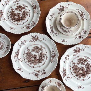 May include: A collection of white ceramic plates, saucers, and teacups with a brown pastoral scene design. The plates have a scalloped edge and floral accents. The teacups and saucers are also white with brown designs.