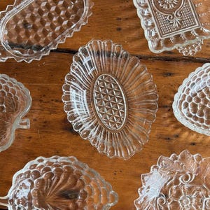 May include: A collection of seven clear pressed glass dishes in various shapes and patterns, including oval, rectangular, and leaf-inspired designs. Each piece features intricate embossed textures, scalloped edges, and geometric motifs.