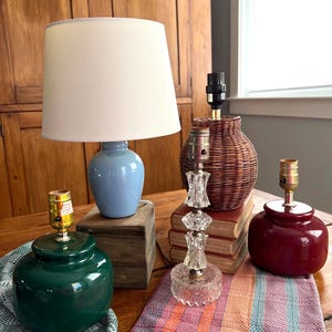 May include: A collection of decorative table lamps in various styles and colors. Includes a blue ceramic lamp with a white shade, a green ceramic lamp, a clear glass lamp, a woven wicker lamp, and a maroon ceramic lamp. The lamps are arranged on a wooden table.