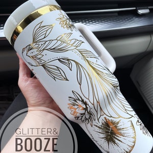 May include: White tumbler with a gold lid and handle. The tumbler has a gold foil design of fish and flowers. The text "Glitter & Booze Shop" is visible on the bottom of the tumbler.