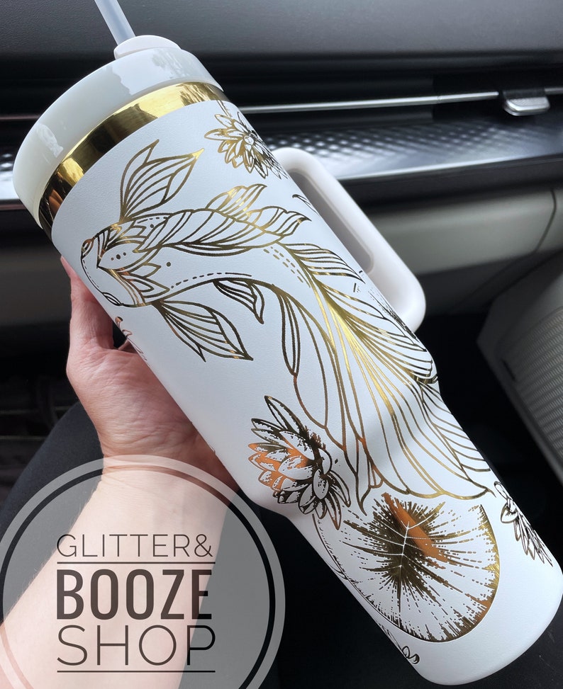 May include: White tumbler with a gold lid and handle. The tumbler has a gold foil design of fish and flowers. The text "Glitter & Booze Shop" is visible on the bottom of the tumbler.