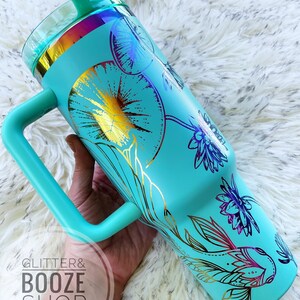 May include: A turquoise stainless steel tumbler with a gold and purple floral design and a fish. The tumbler has a handle and a lid. The tumbler is on a white fuzzy surface.