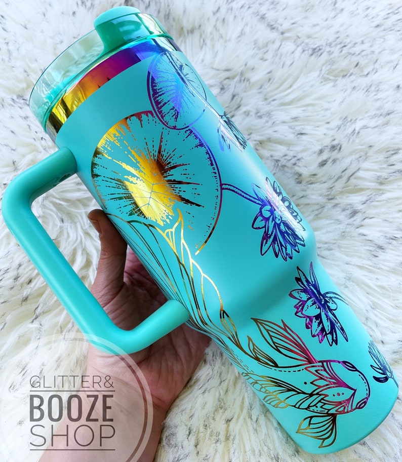May include: A turquoise stainless steel tumbler with a gold and purple floral design and a fish. The tumbler has a handle and a lid. The tumbler is on a white fuzzy surface.