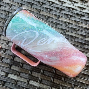 May include: A pink and white marble tumbler with a teal glitter accent and the word "Remarkable" written in white. The tumbler has a handle and is made of stainless steel.