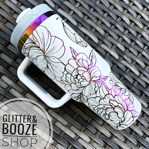 May include: White travel mug with a floral design and a rainbow iridescent lid. The mug has a handle and a black outline of the flowers. The mug is on a woven surface.