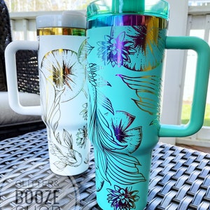 May include: Two stainless steel tumblers with lids and handles. The tumbler on the left is white with gold floral designs. The tumbler on the right is turquoise with gold floral designs. Both tumblers have a glitter and booze shop logo.