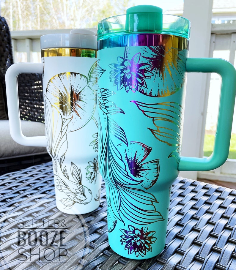 May include: Two stainless steel tumblers with lids and handles. The tumbler on the left is white with gold floral designs. The tumbler on the right is turquoise with gold floral designs. Both tumblers have a glitter and booze shop logo.