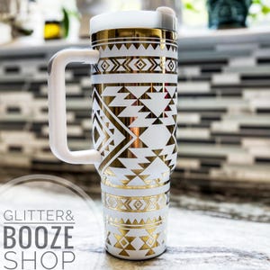 May include: White and gold stainless steel travel mug with a geometric pattern. The mug has a white handle and a lid. The mug is sitting on a white countertop.