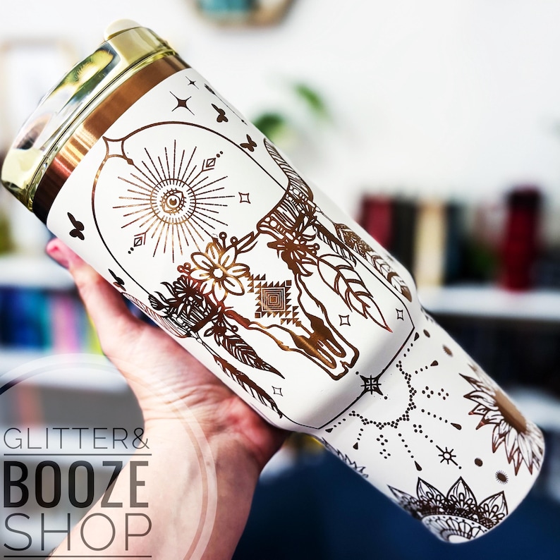 Peut inclure: Un gobelet en acier inoxydable blanc avec un motif m&eacute;tallique dor&eacute; de capteur de r&ecirc;ves avec un cr&acirc;ne de taureau et des accents floraux. Le gobelet a un couvercle dor&eacute;.