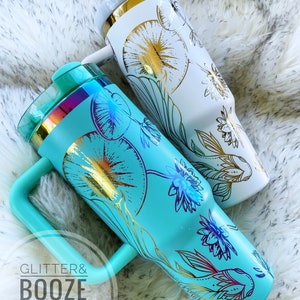 May include: Two stainless steel tumblers with a floral design and gold accents. The tumbler on the left is turquoise blue with a gold fish design. The tumbler on the right is white with a gold floral design. Both tumblers have a handle.