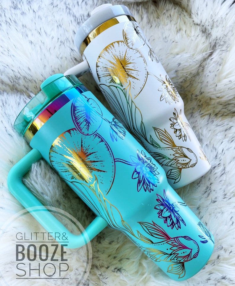 May include: Two stainless steel tumblers with a floral design and gold accents. The tumbler on the left is turquoise blue with a gold fish design. The tumbler on the right is white with a gold floral design. Both tumblers have a handle.