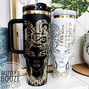 May include: Two stainless steel travel mugs featuring a gold snake and woman design. The mug on the left is black with gold accents. The mug on the right is white with gold accents. Both mugs have a gold rim and a gold handle.