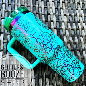 May include: A turquoise tumbler with a black floral design. The tumbler has a handle and a lid. The tumbler is on a woven background.
