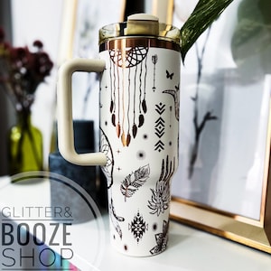 Peut inclure: Un gobelet en acier inoxydable blanc avec un couvercle en cuivre et un motif noir et cuivre repr&eacute;sentant des attrape-r&ecirc;ves, des plumes et des motifs g&eacute;om&eacute;triques. Le gobelet a une poign&eacute;e.