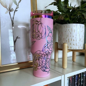 May include: Pink stainless steel tumbler with a gold floral design. The tumbler has a clear lid and a metallic pink finish.