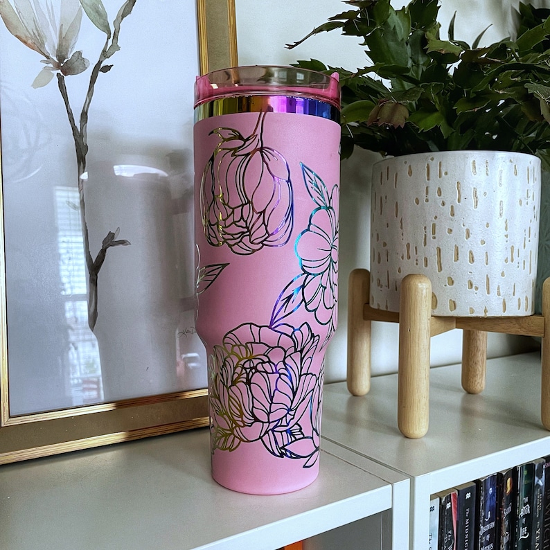 May include: Pink stainless steel tumbler with a gold floral design. The tumbler has a clear lid and a metallic pink finish.
