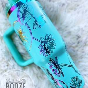 May include: A turquoise blue stainless steel tumbler with a handle. The tumbler has a floral design with gold and iridescent accents. The text "GLITTER & BOOZE SHOP" is visible in the lower left corner of the image.