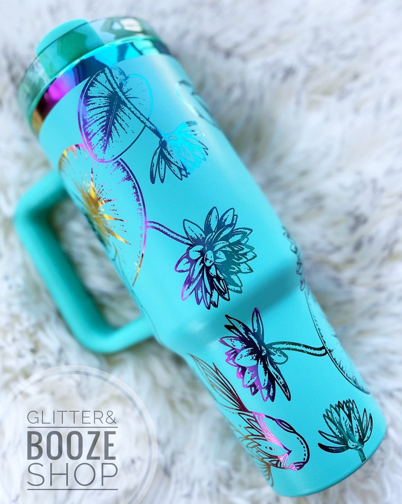 May include: A turquoise blue stainless steel tumbler with a handle. The tumbler has a floral design with gold and iridescent accents. The text "GLITTER & BOOZE SHOP" is visible in the lower left corner of the image.