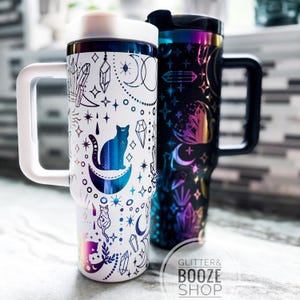 May include: Two insulated travel mugs with handles. One is white with a celestial cat and moon design, the other is black with a colorful crystal and moon design. Both mugs have a white or black handle and lid.