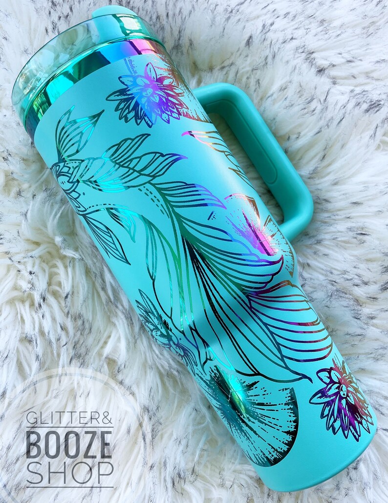 May include: A turquoise stainless steel tumbler with a handle and a floral design. The design is iridescent and features a variety of flowers and leaves. The tumbler is on a white fluffy surface.