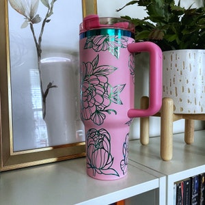 May include: Pink stainless steel tumbler with a floral design and a pink lid and handle. The tumbler is sitting on a white shelf.