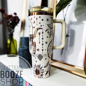 Peut inclure: Mug de voyage blanc avec un motif m&eacute;tallique dor&eacute; repr&eacute;sentant des attrape-r&ecirc;ves, des fl&egrave;ches, des plumes et un tournesol. Le mug est dot&eacute; d'un couvercle et d'une poign&eacute;e.
