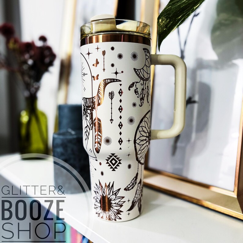 Peut inclure: Mug de voyage blanc avec un motif m&eacute;tallique dor&eacute; repr&eacute;sentant des attrape-r&ecirc;ves, des fl&egrave;ches, des plumes et un tournesol. Le mug est dot&eacute; d'un couvercle et d'une poign&eacute;e.