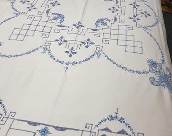 May include: A white tablecloth featuring intricate blue embroidery. The design includes geometric square patterns, delicate floral motifs, and symmetrical scrollwork borders that frame the central area of the fabric.