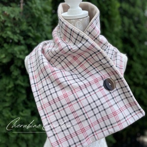 May include: A white, red, and black plaid infinity scarf with a brown button. The scarf is folded in half and is displayed on a white mannequin.