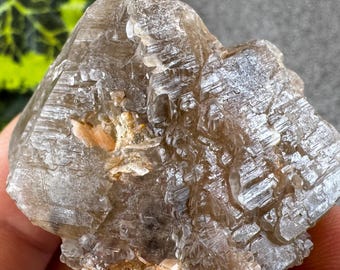 May include: A rough, light brown crystal cluster with a textured surface. The crystal is held in a hand, with a blurred green and yellow plant in the background. The crystal has a natural, earthy appearance.