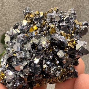 May include: A cluster of metallic pyrite and galena crystals. The crystals display a mix of silver, gold, and black hues, with a sparkling, reflective surface. The mineral specimen is held in a hand, showcasing its natural, raw form.