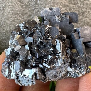 May include: A cluster of dark, metallic-looking crystals with a complex, geometric structure. The crystals are primarily black with reflective surfaces, and some areas show a silver sheen. The overall shape is irregular and rough, with a few small, clear crystals interspersed.