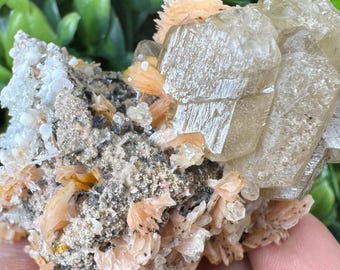 May include: A close-up of a mineral specimen featuring a cluster of translucent, light-brown crystals. The crystals are surrounded by smaller, peach-colored formations and sit on a matrix of gray and white rock. A purple flower is visible in the background.