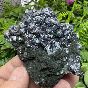 Puede incluir: Un espécimen mineral plateado y negro con una superficie rugosa y texturizada. El mineral tiene un brillo metálico y se coloca sobre un fondo verde.